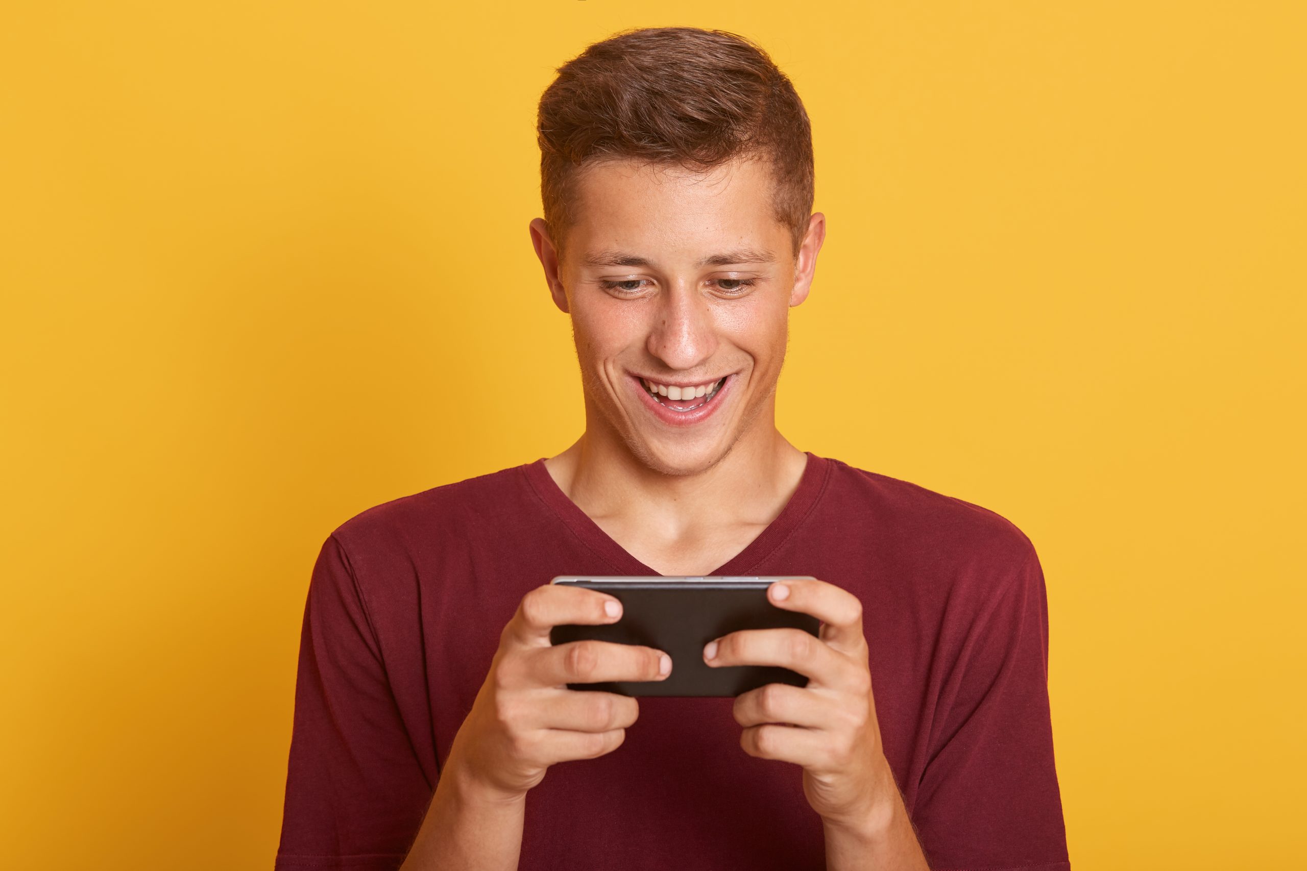 Smiling young man playing game on smartphone isolated over yellow background, looks happy and concentrated, looking smiling at his device's screen, spending spare time indoor, guy holding mobile phone